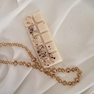 May include: A rectangular, off-white wax melt bar with glitter accents, resting on a white fabric. A gold beaded necklace is draped near the wax melt. The wax melt is divided into ten squares.