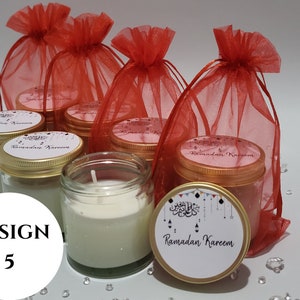 May include: A set of six small glass jars with gold lids filled with white candles. The jars are decorated with a white label that says "Ramadan Kareem" in Arabic script. The jars are arranged in a group on a white surface with red mesh bags behind them.
