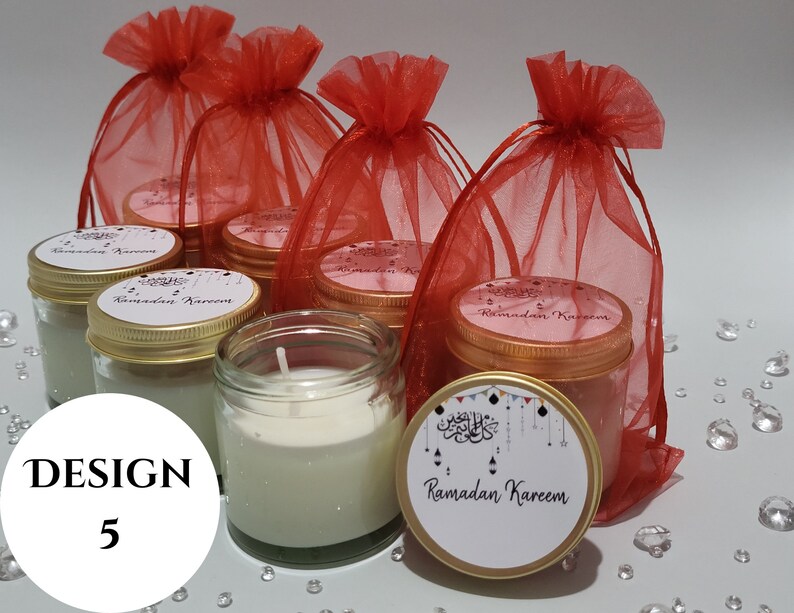 May include: A set of six small glass jars with gold lids filled with white candles. The jars are decorated with a white label that says "Ramadan Kareem" in Arabic script. The jars are arranged in a group on a white surface with red mesh bags behind them.