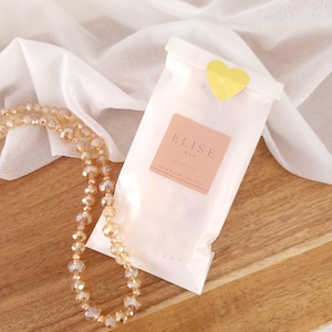 May include: A white wax melt bag with a yellow heart-shaped sticker, a pink label with the text "ELISE WAX", and a beaded necklace. The bag is on a wooden surface with a white fabric in the background.