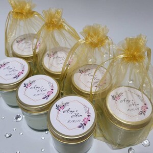 Puede incluir: Conjunto de seis peque&ntilde;os frascos de vidrio con tapas doradas y etiquetas personalizadas. Cada frasco tiene una etiqueta blanca con un dise&ntilde;o floral y el texto "Amy & Hen 01/06/23". Los frascos est&aacute;n empaquetados en bolsas individuales de organza dorada.