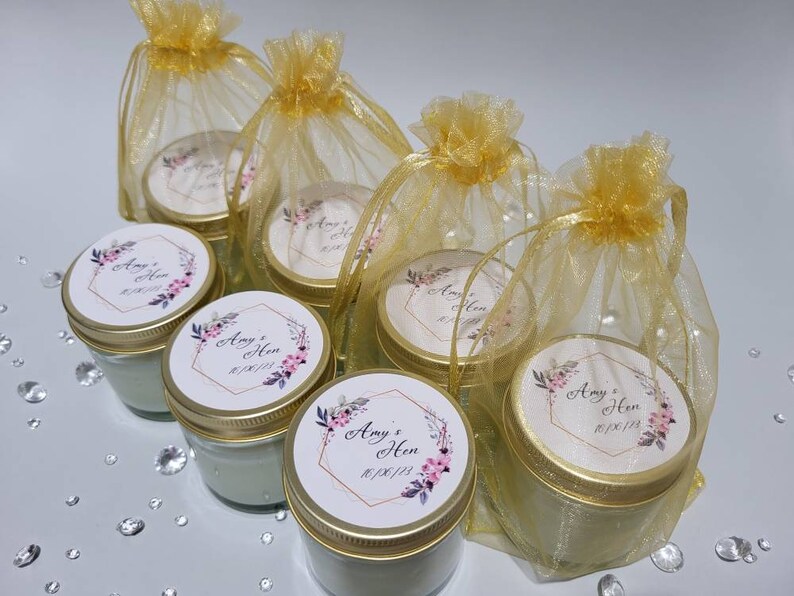 Puede incluir: Conjunto de seis peque&ntilde;os frascos de vidrio con tapas doradas y etiquetas personalizadas. Cada frasco tiene una etiqueta blanca con un dise&ntilde;o floral y el texto "Amy & Hen 01/06/23". Los frascos est&aacute;n empaquetados en bolsas individuales de organza dorada.