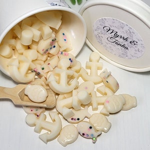 May include: A white paper cup filled with small, white wax melts shaped like anchors, fish, and other nautical shapes. The wax melts are sprinkled with colorful glitter. The cup is labeled "Myrrh & Tonka".