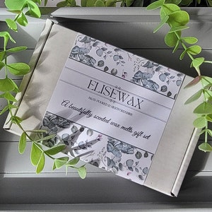 K&ouml;nnte beinhalten: Ein Geschenkset mit Duftwachs-Schmelzen in einer wei&szlig;en Schachtel mit floralem Design, auf einem grauen Tablett mit gr&uuml;nem Laub. Auf der Schachtel steht "ELISEWAX" und "A beautifully scented wax melts gift set."