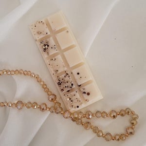 May include: A rectangular wax melt bar with ten sections, off-white in color, and speckled with gold and black glitter. A beaded necklace with gold-colored beads is draped below the wax melt bar. The background is a white, silky fabric.