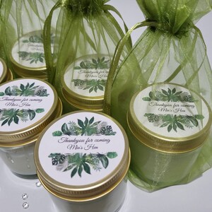 Puede incluir: Peque&ntilde;os frascos de vidrio con tapas doradas y etiquetas verdes y blancas que dicen "Thankyou for coming Mia's Hen". Los frascos est&aacute;n en bolsas de organza verde.
