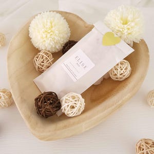 Könnte beinhalten: Eine herzförmige Holzschale enthält eine weiße Papierverpackung mit dem Text "ELISE WAX" und einem gelben herzförmigen Aufkleber. Dekorative Kugeln und zwei cremefarbene Blumen umgeben die Verpackung.