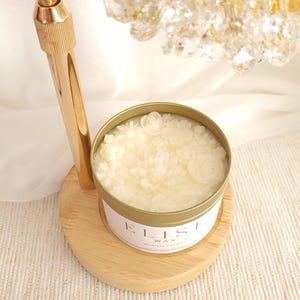 Könnte beinhalten: Eine cremefarbene Kerze in einer goldenen Dose mit floralem Design. Die Kerze steht auf einem Holzsockel, mit dem Text "ELISE WAX" auf dem Etikett. Ein goldfarbener Metallständer ist im Hintergrund.