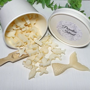 May include: Off-white wax melts in various sea creature shapes, including starfish, dolphins, and a mermaid tail, spilling from a white container. The container lid has a label that reads "Paradise Island". A small wooden scoop is also present.