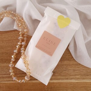 May include: A necklace with champagne-colored beads and a white paper bag sealed with a yellow heart sticker. The bag has a label that reads "ELISE WAX" and "Hand Poured | Est. 2021". The background includes a white cloth and a wooden surface.