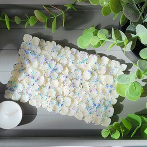 K&ouml;nnte beinhalten: Ein rechteckiger, cremefarbener Wachsschmelz in Form eines Blumenclusters, &uuml;bers&auml;t mit blauem und violettem Glitzer. Daneben steht eine kleine, wei&szlig;e Teelichtkerze auf einem grauen Tablett, mit gr&uuml;nem Laub im Hintergrund.