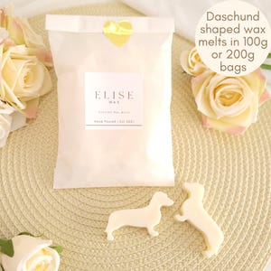Könnte beinhalten: Ein weißer Papiersack mit dem Text "ELISE WAX" und "Hand Poured | Est 2021". Der Beutel ist mit einem goldenen Herzaufkleber verschlossen. Zwei dackelformige Wachsschmelzen befinden sich vor dem Beutel. Der Text "Daschund shaped wax melts in 100g or 200g bags" ist ebenfalls sichtbar.