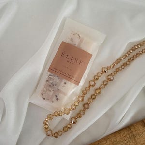May include: A package of Elise Wax melts with a brown label and the text "Hand Poured | Est 2021". A gold-colored beaded necklace is next to the wax melts. The background is a white, draped fabric.