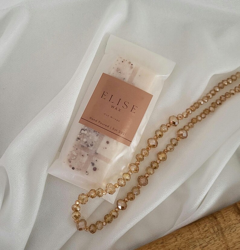 May include: A package of Elise Wax melts with a brown label and the text "Hand Poured | Est 2021". A gold-colored beaded necklace is next to the wax melts. The background is a white, draped fabric.