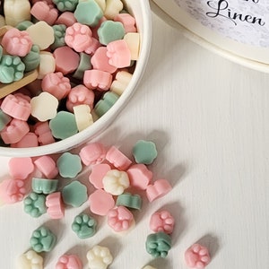 May include: A white bowl filled with pastel pink, green, and white paw print shaped wax melts. Some of the melts are scattered on a white surface.