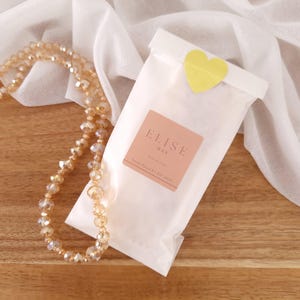 May include: A white wax melt bag with a yellow heart sticker, a peach-colored label with the text "ELISE WAX", and a beaded necklace. The bag is on a wooden surface with a white cloth in the background.