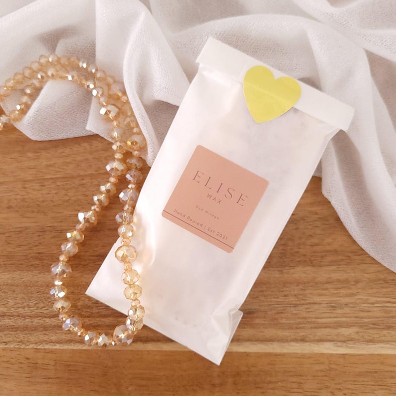 May include: A white wax melt bag with a yellow heart sticker, a peach-colored label with the text "ELISE WAX", and a beaded necklace. The bag is on a wooden surface with a white cloth in the background.
