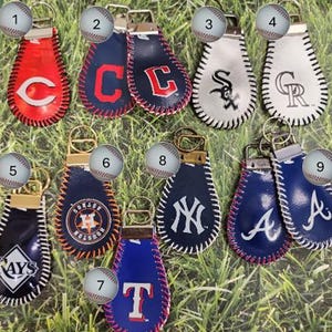 May include: A collection of baseball-themed keychains in various colors, featuring team logos and stitched edges. The keychains are made of leather and have a metal keyring attachment. Logos include the Astros, Yankees, and Rays.