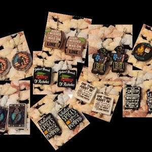 May include: A collection of novelty earrings with various designs. Some earrings feature text like "Sorry I missed your call, I was on the other line," and "Jolliest Bunch of Assholes." Other designs include a campfire, a soldier, and a deck of cards.