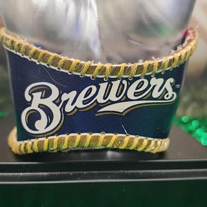May include: A navy blue and white Brewers logo is displayed on a decorative item. The logo is set against a navy blue background, with a yellow stitched border. The item is on a black base.