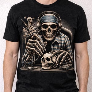 May include: Dark gray t-shirt featuring a detailed illustration of a skeleton wearing headphones and a bandana, tattooing a skull. The design includes a tattoo machine and a second skull on a table.
