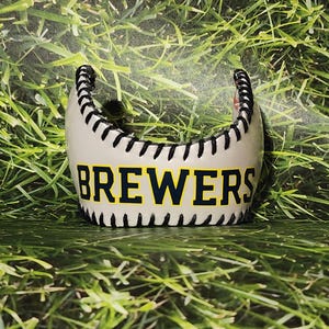 May include: A baseball-themed decorative item with the word "BREWERS" in navy blue and yellow lettering. The item is white with black stitching, resembling a baseball. The background is green grass.