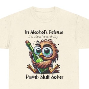 May include: Cream-colored t-shirt featuring a cartoon owl holding a green bottle. The owl has brown and tan feathers, large green eyes, and yellow feet. Text on the shirt reads "In Alcohol's Defense I've Done Some Pretty Dumb Stuff Sober."