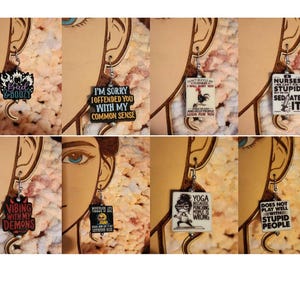 May include: A collection of humorous earrings with various phrases and graphics. The earrings feature text like "Bad & Boozy", "I'm Sorry I Offended You", and "Nurses...Sedate It". Each earring is rectangular or uniquely shaped, with a silver hook.