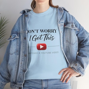 May include: A light blue t-shirt with the text "DON'T WORRY I Got This" and a red YouTube play button graphic. Below the graphic, the text reads "WATCHED A YOUTUBE VIDEO". The shirt is worn with a denim jacket.