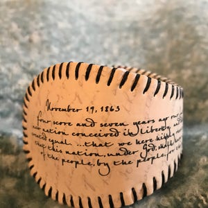 May include: A white leather bracelet with black stitching and the text "November 19, 1863 four score and seven years ago our nation conceived in Liberty and that we here highly resolve that this nation, under God, shall have a new birth of freedom - and that government of the people, by the people, for the people, shall not perish from this earth."