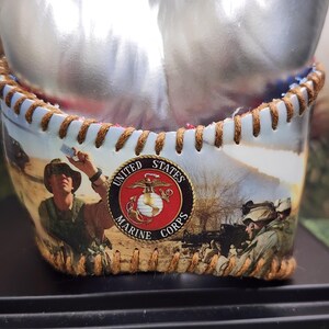 May include: A decorative container with a stitched leather rim and a printed image of U.S. Marines in action. The container features the United States Marine Corps emblem.