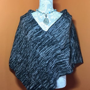 May include: Black and white knit poncho with a v-neck. The poncho is draped over a mannequin wearing a necklace with a silver pendant.