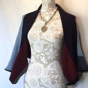 May include: A blue, burgundy, and navy blue knitted shrug with a draped design. The shrug is displayed on a mannequin wearing a necklace with a large teardrop pendant.