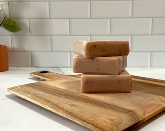 May include: Three bars of natural soap stacked on a wooden cutting board. The soap is a light brown color with flecks of red and white.