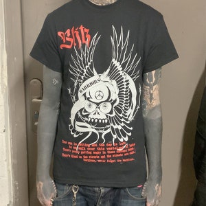 以下が含まれることがあります： 白い骸骨と翼のグラフィックが描かれた黒いTシャツ。骸骨は「WARRIORS」と書かれたヘルメットをかぶっています。Tシャツのテキストは次のとおりです。「Your sun is setting and the day is late. Is we walk over this wasteland of hate. There's you getting angry in these darkest hours. There's blood on the streets and the streets are ours. Warriors, never forget the Warriors.」