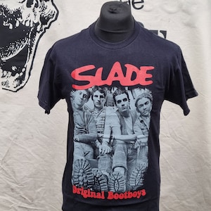 Puede incluir: Camiseta negra con un gráfico de banda que presenta una foto de cuatro hombres. El nombre de la banda "SLADE" está impreso en rojo sobre la foto. El texto "Original Bootboys" está impreso en rojo debajo de la foto.