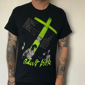 May include: Black t-shirt with a green cross graphic and the text "Adult Books" in white. The shirt features a band of four people in black and white.
