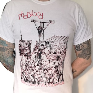 Puede incluir: Camiseta blanca con una imagen en blanco y negro de una persona en una cruz rodeada de gente. El texto "theBlood" está impreso en rojo sobre la imagen. El texto "False Gestures for a Devious Public" está impreso en rojo debajo de la imagen.