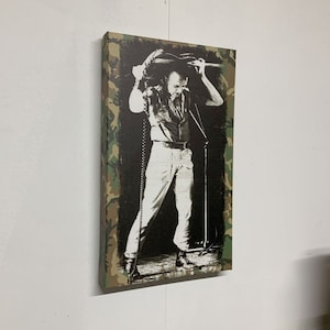 Op de afbeelding: Een zwart-witfoto van een muzikant die een gitaar boven zijn hoofd houdt. De foto is afgedrukt op een canvas met een camouflage-patroonrand.