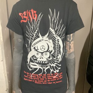 以下が含まれることがあります： 白い翼と有刺鉄線が付いた骸骨のグラフィックが描かれた黒いTシャツ。骸骨には「WARRIORS」の文字が書かれている。「Your sun is setting and the day is late. There's blood on the streets and the streets are cursed. Warriors, never forget the Warriors.」というテキストがシャツにプリントされている。