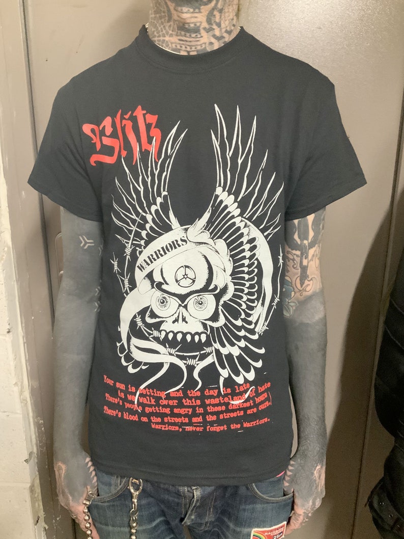 以下が含まれることがあります： 白い翼と有刺鉄線が付いた骸骨のグラフィックが描かれた黒いTシャツ。骸骨には「WARRIORS」の文字が書かれている。「Your sun is setting and the day is late. There's blood on the streets and the streets are cursed. Warriors, never forget the Warriors.」というテキストがシャツにプリントされている。