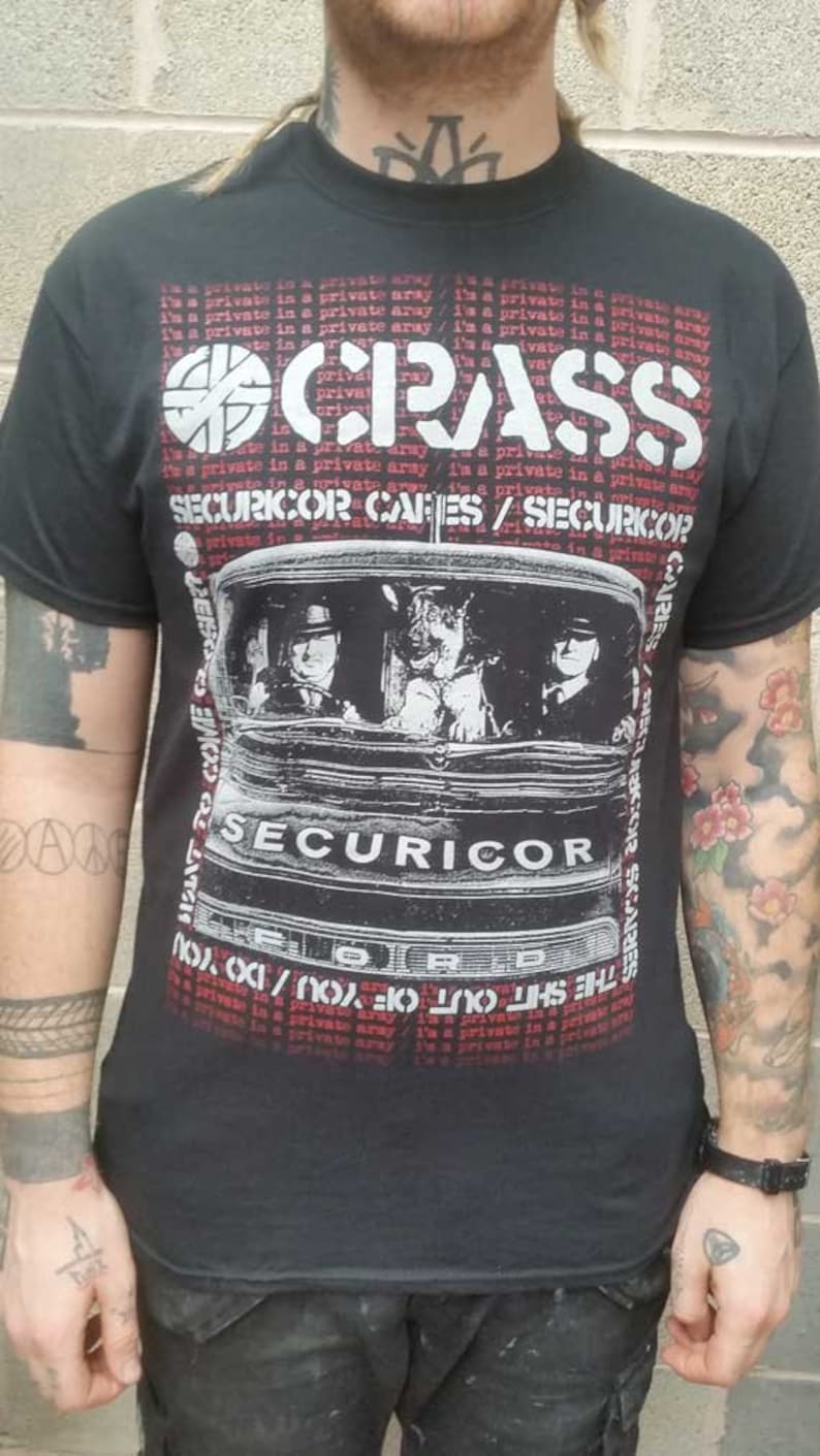 May include: Black t-shirt featuring a white graphic of a van with three people inside. The text "SECURICOR CARES / SECURICOR" is printed in red above the van. The text "I'm a private in a private array / I'm a private in a private array" is printed in red above and below the graphic.