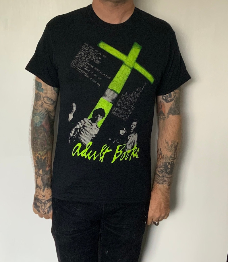 May include: Black t-shirt with a green cross graphic and the text "Adult Books" in white. The shirt features a band of four people in black and white.