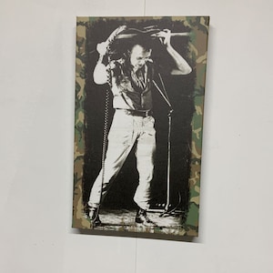 Op de afbeelding: Zwart-witfoto van een muzikant die een gitaar boven zijn hoofd houdt. De foto is afgedrukt op een canvas met een camouflage-rand.