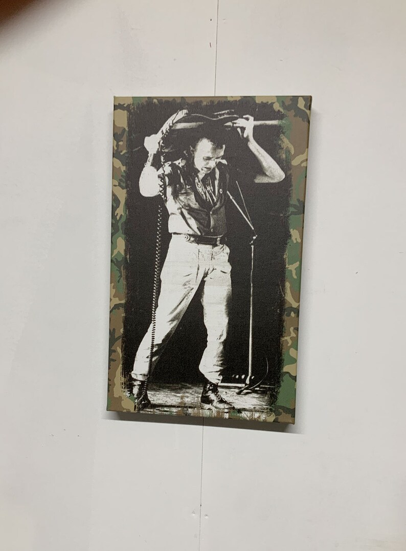 Op de afbeelding: Zwart-witfoto van een muzikant die een gitaar boven zijn hoofd houdt. De foto is afgedrukt op een canvas met een camouflage-rand.