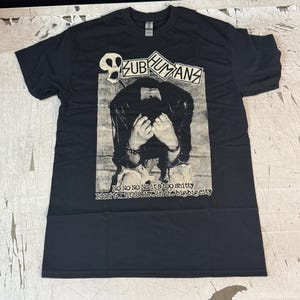 Puede incluir: Camiseta negra con un gráfico. El gráfico incluye el nombre de la banda "SUBHUMANS" y una imagen en blanco y negro de una persona con las manos cubriendo su rostro. El texto debajo de la imagen dice "NO NO NO NO it's too shitty I don't want to live in the big big city".