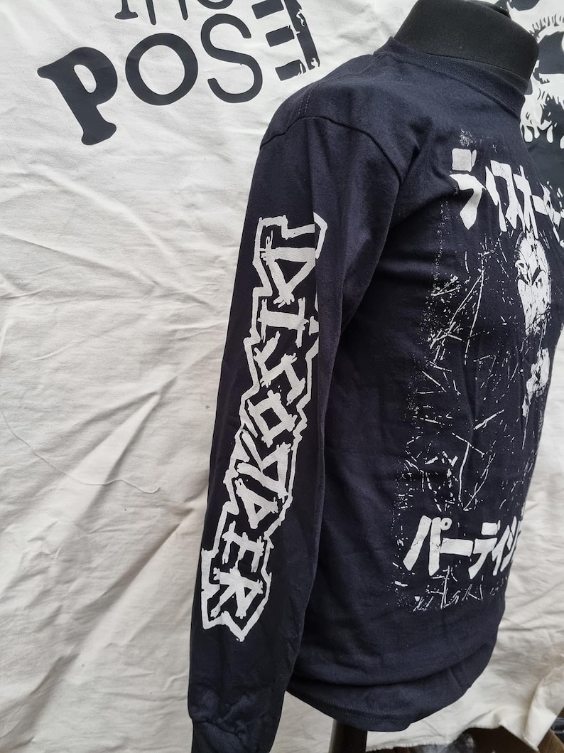 K&ouml;nnte beinhalten: Schwarzes Langarmshirt mit wei&szlig;em Text auf den &Auml;rmeln und dem R&uuml;cken. Der Text auf den &Auml;rmeln lautet "DISCHARGE" und der Text auf dem R&uuml;cken lautet "Discharge" in japanischen Schriftzeichen und "パーディション" in japanischen Schriftzeichen.