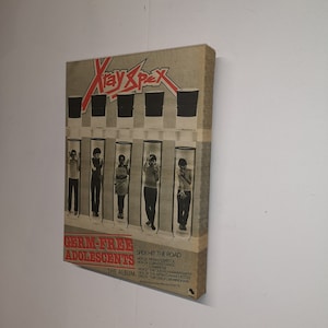 Pu&ograve; includere: Una copertina di un album in vinile vintage con una band chiamata "X-Ray Spex" con il titolo "Germ-Free Adolescents." La copertina presenta sei persone in provette, con il nome della band in lettere rosse e nere.