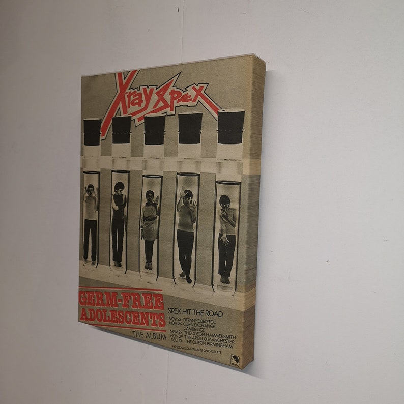 Pu&ograve; includere: Una copertina di un album in vinile vintage con una band chiamata "X-Ray Spex" con il titolo "Germ-Free Adolescents." La copertina presenta sei persone in provette, con il nome della band in lettere rosse e nere.