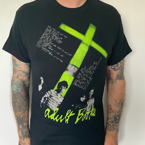 May include: Black t-shirt with a green cross graphic and the text "adult Books" printed on the front. The shirt features a band of lyrics printed on the side.